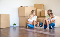 Smiling couple sitting with two daughters and looking at each other blonde mother and middle aged father with their children relaxing near carton boxes mortgage relocation and moving day 820955581